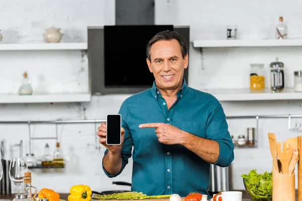 Sorrindo bonito homem de meia-idade apontando no smartphone com tela em branco na cozinha — Fotografia de Stock