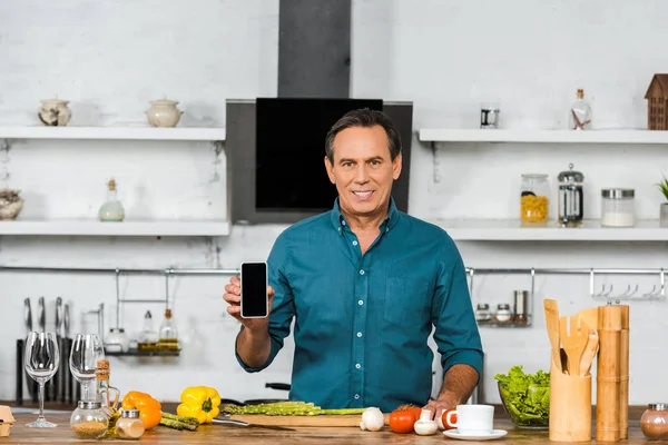 Sourire bel homme d'âge moyen tenant smartphone avec écran blanc dans la cuisine — Photo de stock