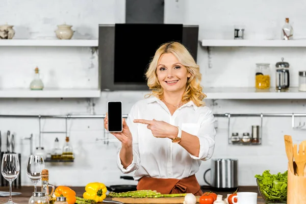 Attractive middle aged woman pointing on smartphone with blank screen in kitchen — Stock Photo