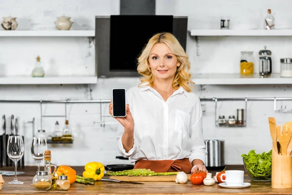 Attraente donna di mezza età che mostra smartphone con schermo bianco in cucina — Foto stock