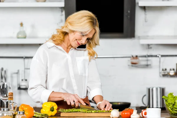 Attractive middle aged woman talking by smartphone while cooking in kitchen — Stock Photo