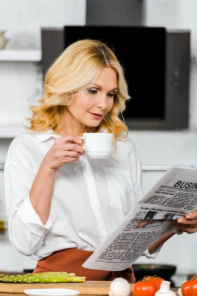 Schöne Frau mittleren Alters hält eine Tasse Tee in der Küche und liest Zeitung — Stockfoto