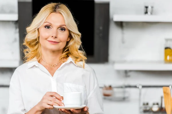 Atractiva mujer de mediana edad sosteniendo taza de té en la cocina y mirando a la cámara - foto de stock