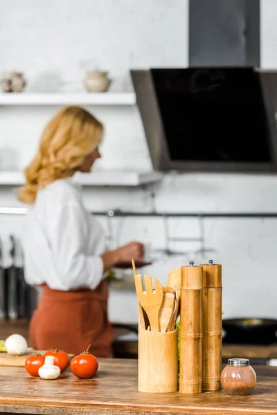 Enfoque selectivo de la mujer de mediana edad sosteniendo la taza de té en la cocina, verduras y utensilios de madera en la mesa - foto de stock