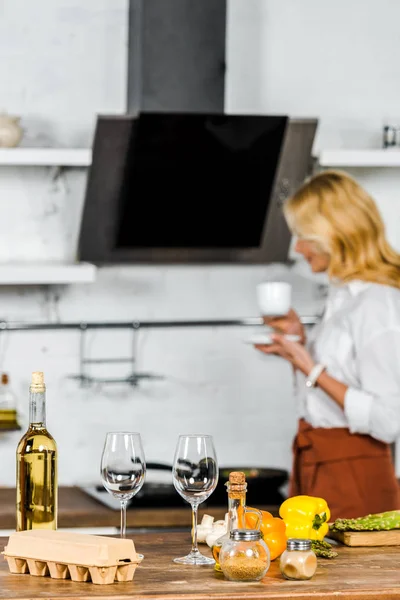 Selective focus of middle aged woman holding cup of tea in kitchen, vegetables and wine on tabletop — Stock Photo