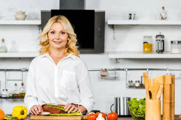 Smiling beautiful middle aged woman cutting vegetables in kitchen and looking at camera — Stock Photo