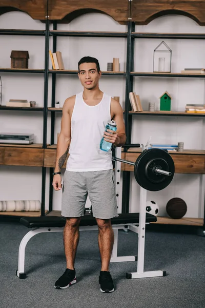 Guapo bi-racial hombre sosteniendo deporte botella y de pie cerca de barbell - foto de stock