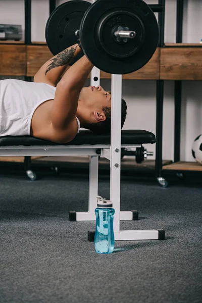 Tattooed mixed race man workout with barbell near sport bottle — Stock Photo