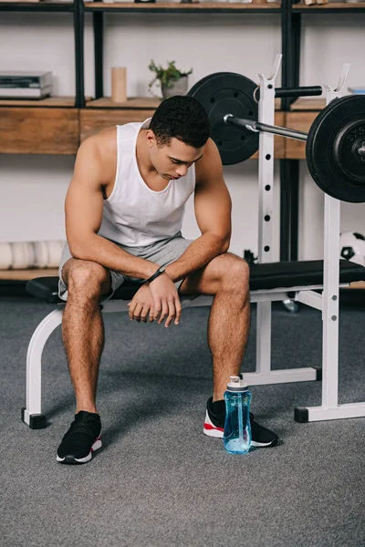 Hombre de raza mixta sentado en ropa deportiva y mirando botella de deporte - foto de stock