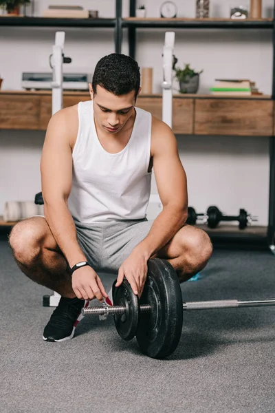 Mixed race man sitting near barbell and putting heavy disk — Stock Photo