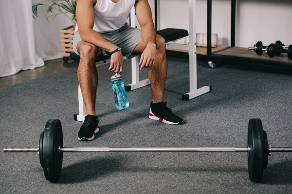 Cropped view of man sitting near barbell and holding sport bottle — Stock Photo