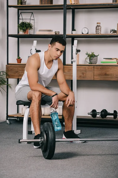 Thoughtful bi-racial man sitting near barbell and holding sport bottle — Stock Photo