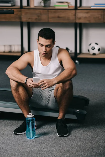 Deportista bi-racial sentado cerca de la botella de deporte y el uso de teléfono inteligente - foto de stock