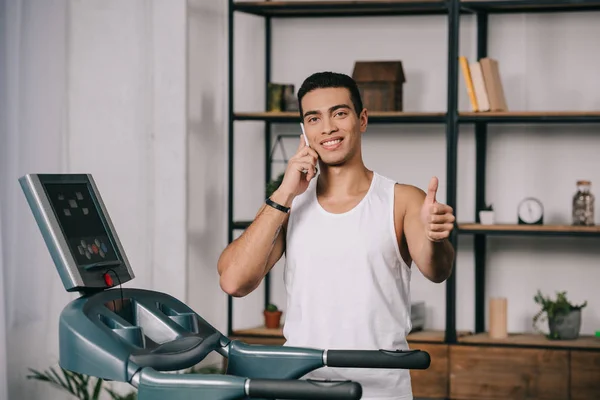 Sonriente deportista bi-racial hablando en smartphone y mostrando el pulgar hacia arriba - foto de stock
