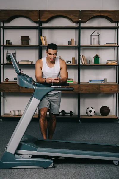 Strong mixed race man using smartphone near treadmill — Stock Photo