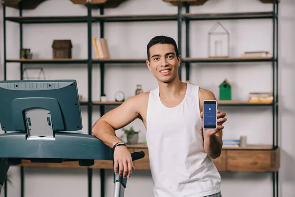 Cheerful bi-racial man holding smartphone with facebook app on screen — Stock Photo