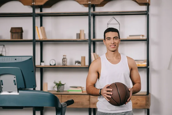 Sonriente hombre de raza mixta celebración de baloncesto en sala de estar - foto de stock