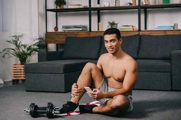 Alegre hombre bi-racial sentado en el suelo cerca de pesas mientras se utiliza el teléfono inteligente en la sala de estar - foto de stock