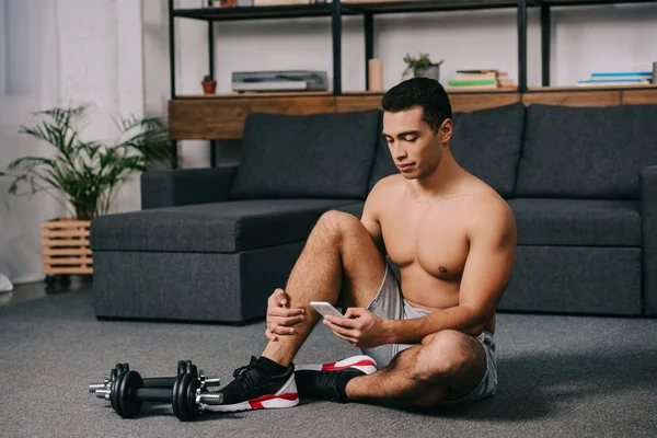 Bi-racial man sitting on floor near dumbbells while using smartphone in living room — Stock Photo