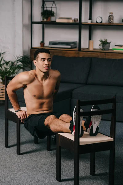Mixed race man exercising on chairs in home gym — Stock Photo
