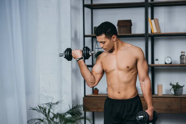 Handsome bi-racial man looking at muscles while exercising with dumbbells in home gym — Stock Photo