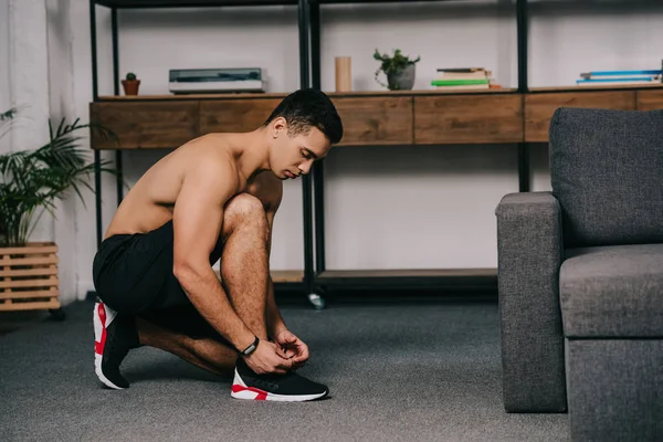 Musculoso bi-racial hombre atando cordones en zapatilla de deporte en la sala de estar - foto de stock