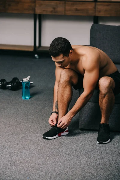 Hombre bi-racial fuerte atando cordones de zapatos cerca de pesas y botella de deporte en la sala de estar - foto de stock