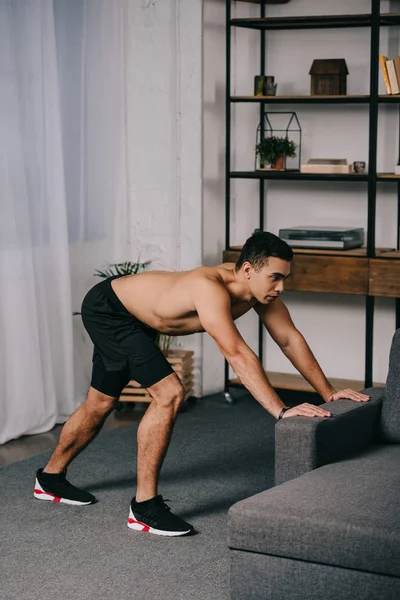 Hombre de raza mixta fuerte haciendo ejercicio cerca del sofá en la sala de estar - foto de stock