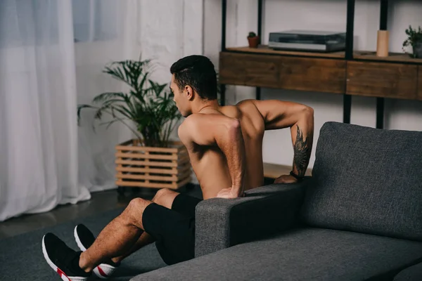 Entrenamiento de hombre de raza mixta tatuado cerca del sofá en la sala de estar - foto de stock