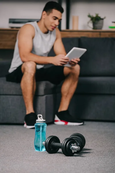 Enfoque selectivo de la botella de deporte cerca de pesas con el hombre bi-racial sosteniendo tableta digital en el fondo - foto de stock