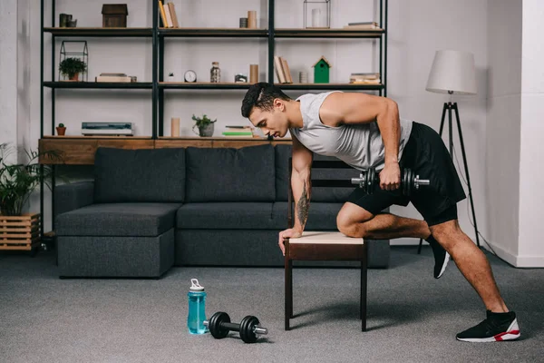 Strong bi-racial man doing exercise with dumbbell on chair in living room — Stock Photo