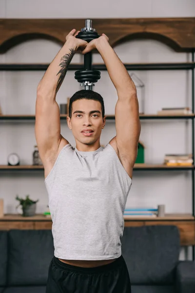 Bonito bi-racial homem de pé no sportswear e segurando dumbbell sobre cabeça — Fotografia de Stock