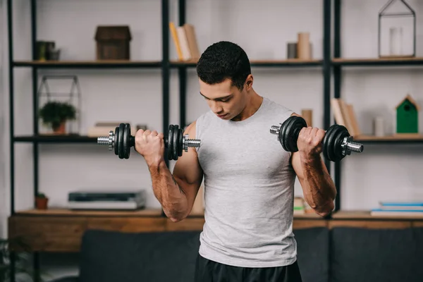 Fuerte mestizo hombre entrenamiento con mancuernas en sala de estar - foto de stock