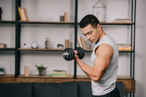 Guapo hombre de raza mixta mirando los músculos en la mano mientras hace ejercicio con la mancuerna en la sala de estar - foto de stock