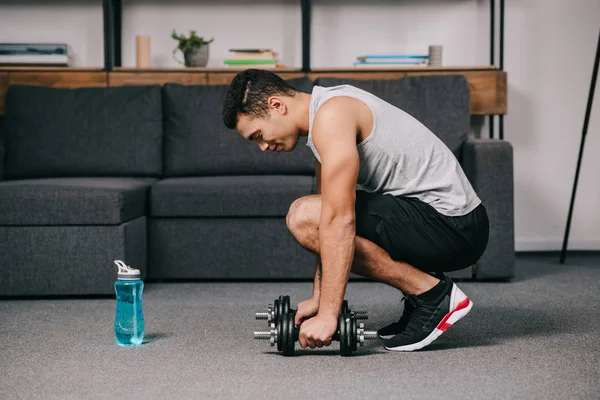 Handsome mixed race sportsman putting dumbbells on floor in living room — Stock Photo