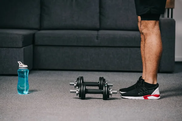 Cropped view of sportsman standing near dumbbells and sport bottle with water in living room — Stock Photo