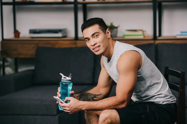 Smiling mixed race man holding sport bottle and smartphone while sitting on chair in living room — Stock Photo