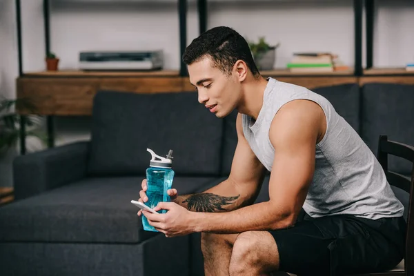 Hombre de raza mixta con tatuaje celebración de botella deportiva y el uso de teléfono inteligente mientras está sentado en la silla en la sala de estar - foto de stock