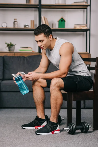 Hombre de raza mixta sosteniendo la botella deportiva y el uso de teléfono inteligente mientras está sentado en la silla en la sala de estar - foto de stock