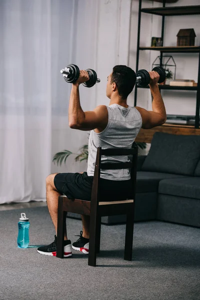 Musculoso hombre de raza mixta sosteniendo pesadas mancuernas mientras está sentado en la silla en el apartamento - foto de stock