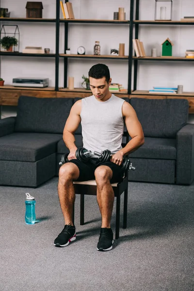 Mixed race athlete sitting with dumbbells on chair in home gym — Stock Photo