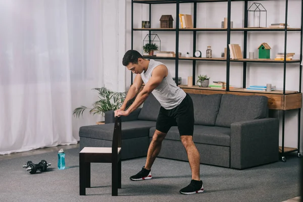 Мускулистый спортсмен смешанной расы, использующий стул для тренировок в гостиной — стоковое фото