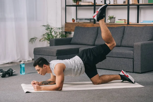 Bel athlète de course mixte avec tatouage à la main faisant de l'exercice sur les jambes sur tapis de fitness dans le salon — Photo de stock