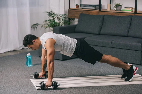 Uomo bi-razziale facendo esercizio plancia con manubri sul tappeto fitness in soggiorno — Foto stock