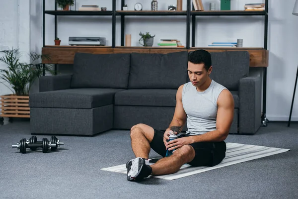 Uomo bi-razziale seduto con bottiglia di sport sul tappeto fitness in soggiorno — Foto stock