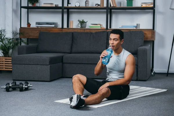 Guapo bi-racial hombre beber agua de deporte botella mientras sentado en fitness mat - foto de stock