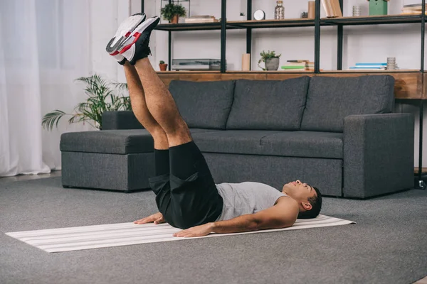 Hombre bi-racial acostado en la alfombra de fitness y haciendo ejercicio en ropa deportiva - foto de stock