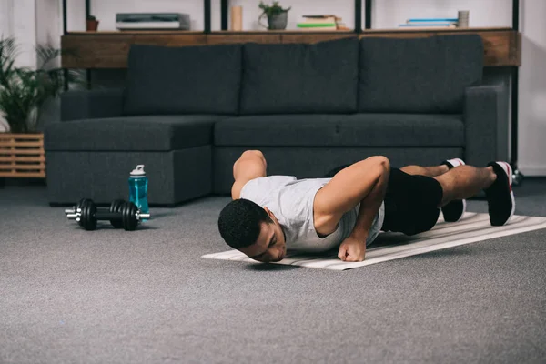 Hombre bi-racial acostado en la alfombra de fitness y haciendo ejercicio en la sala de estar - foto de stock