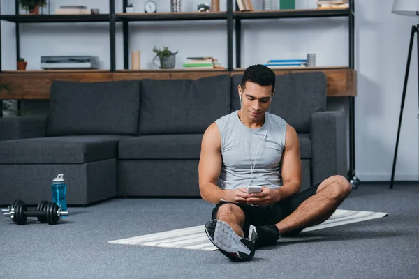 Bi-racial homem segurando smartphone e ouvir música em fones de ouvido no tapete de fitness — Fotografia de Stock