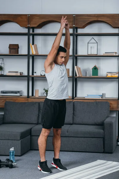 Guapo bi-racial hombre de pie cerca de la alfombra de fitness y haciendo ejercicio - foto de stock